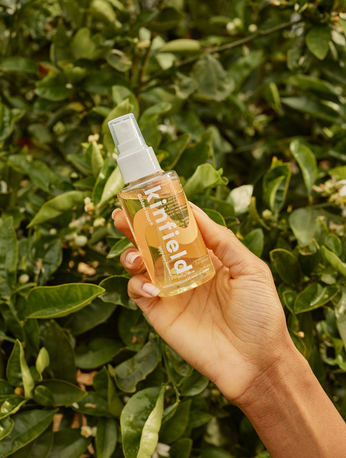 A hand holding a Kinfield Golden Hour™ spray bottle against a backdrop of lush green leaves showcases a clean bug spray solution infused with Indonesian citronella.