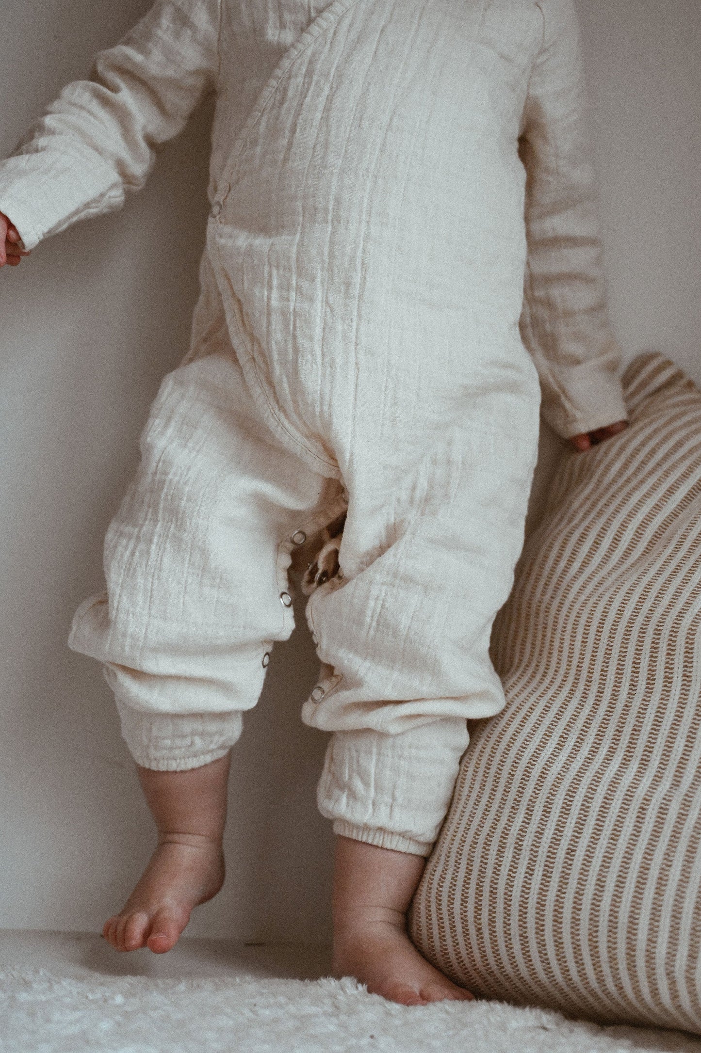 A baby wearing the cozy Après Organic Wrap Undyed from cabane childrenswear's layette collection stands on a soft surface, one hand resting on a striped cushion. Their feet are bare, and the wrap's buttons and texture are visible.