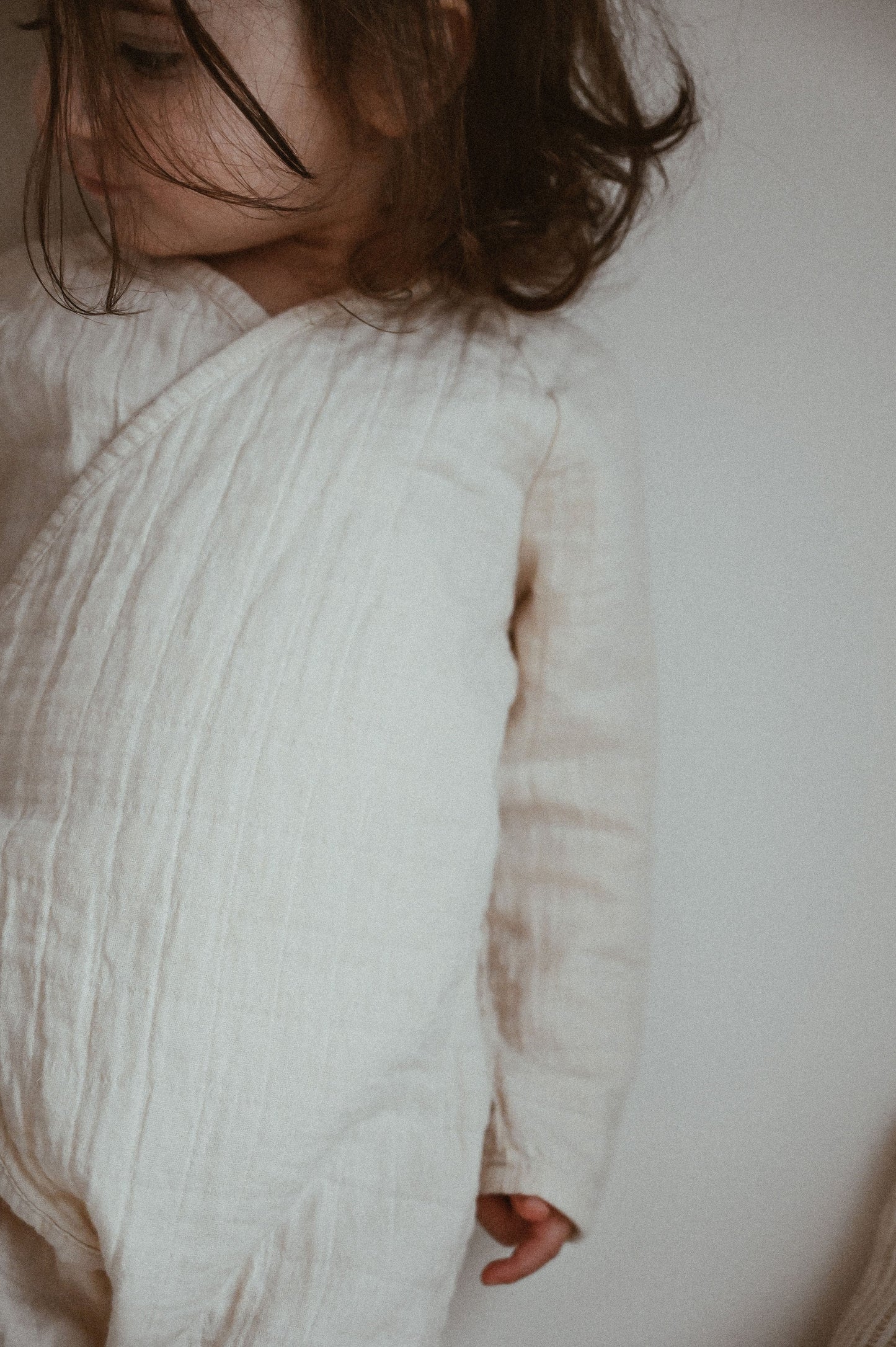 A young child dressed in the Après Organic Wrap Undyed from cabane childrenswear stands slightly turned away, with dark hair partially covering their face.