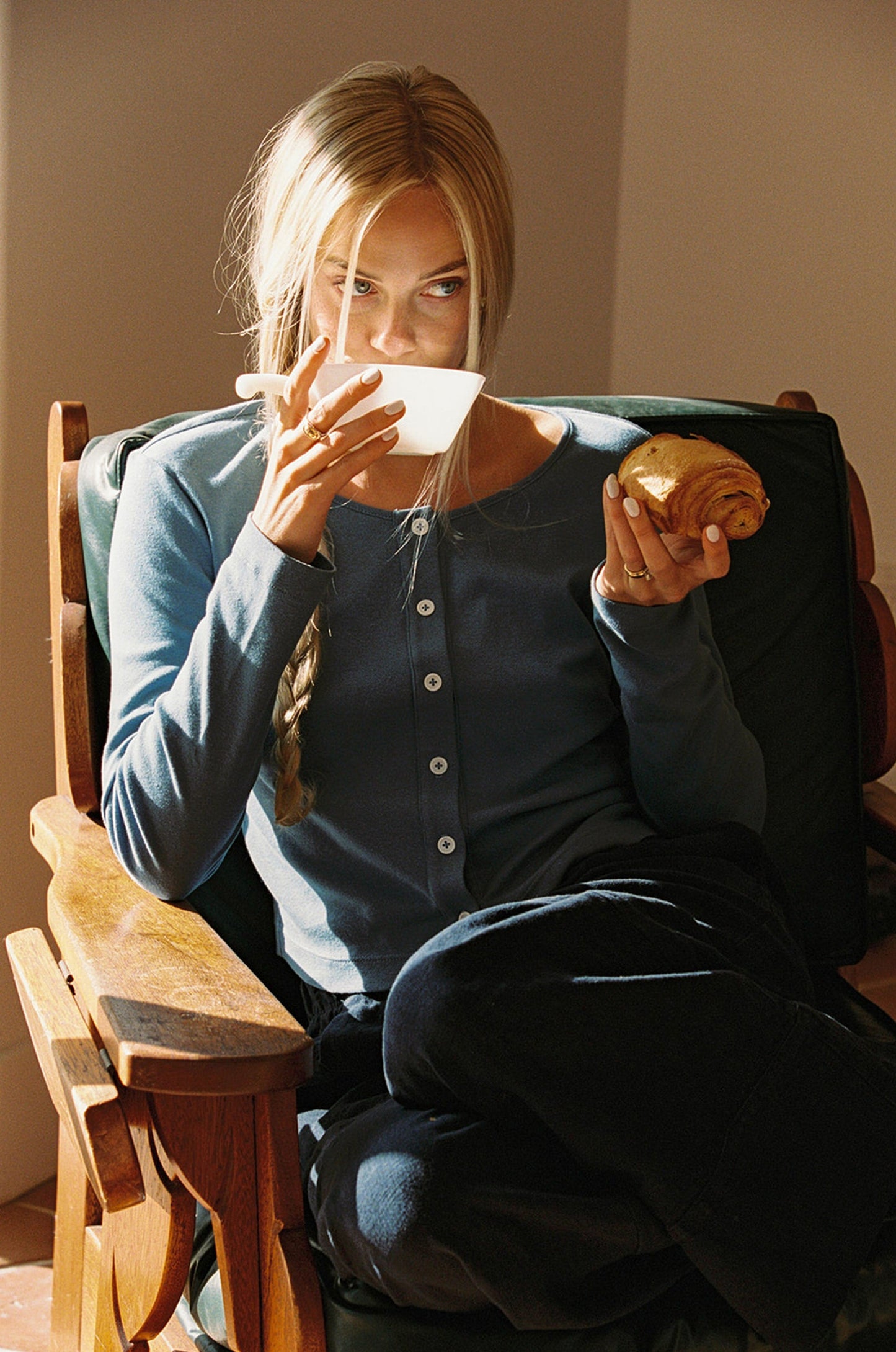 A person with long blonde hair sits in a wooden chair, enjoying a croissant and cup. They wear an Organic Rib Cardigan by LA Relaxed and dark pants, as sunlight casts warm shadows across the scene.