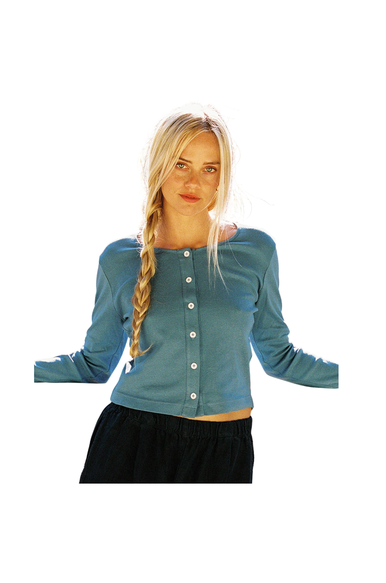 A person with long blonde hair in a braid wears the LA Relaxed Organic Rib Cardigan, featuring pearlescent buttons, paired with dark pants. They stand with arms slightly out to the sides against a completely black background.
