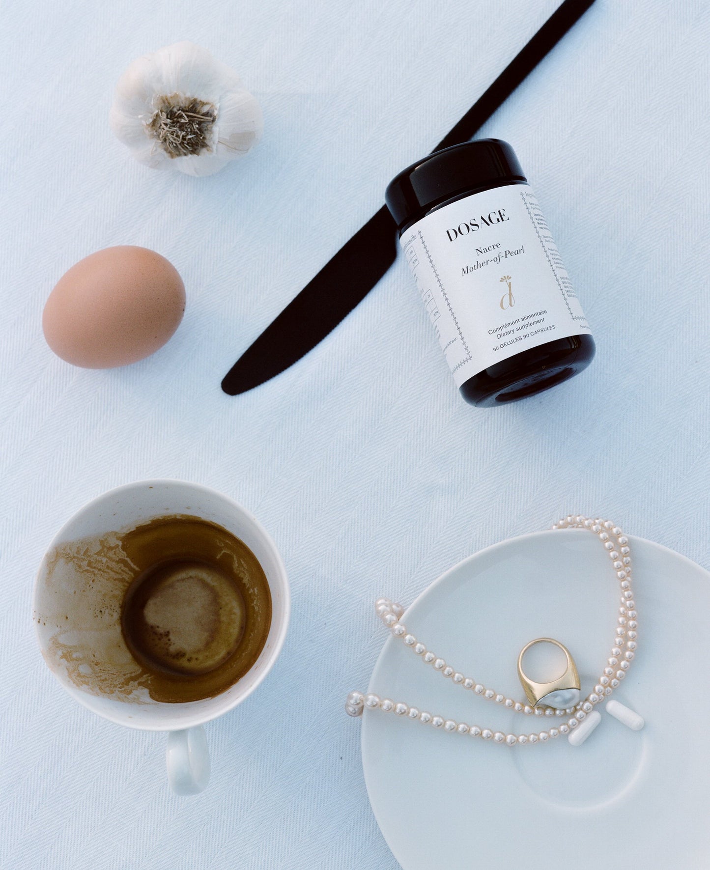 A flat lay photo featuring a garlic bulb, a brown egg, a black knife, a jar labeled "DOSAGE Morning Pearl Mushroom Powder," a saucer with coffee residue in a teacup, and a saucer with Mother-of-Pearl extract adorned with a pearl necklace and gold ring against a white background.