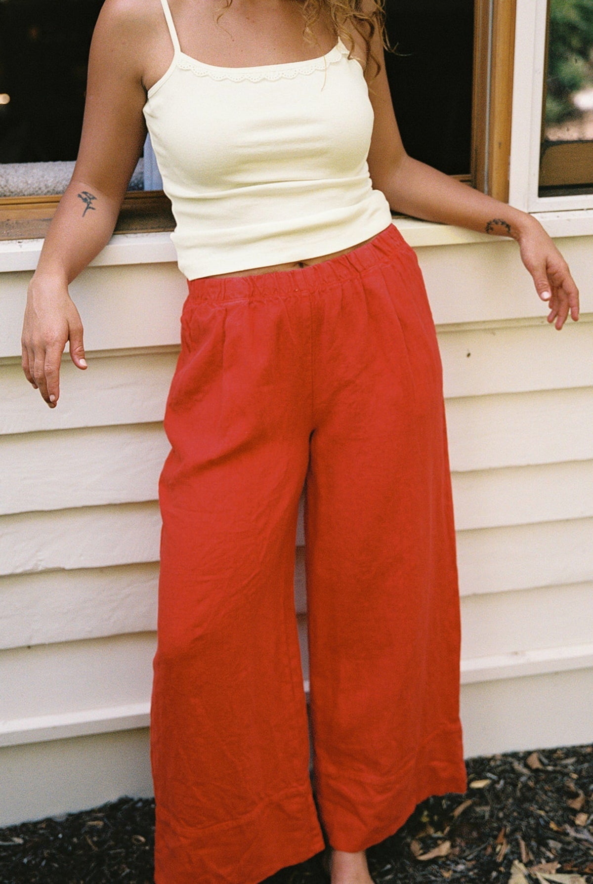 Cream -
A person stands outside in front of a white wall wearing the LA RELAXED Frill Camisole in pale yellow and loose red pants. Their face is not visible, and one arm rests on a window frame.
