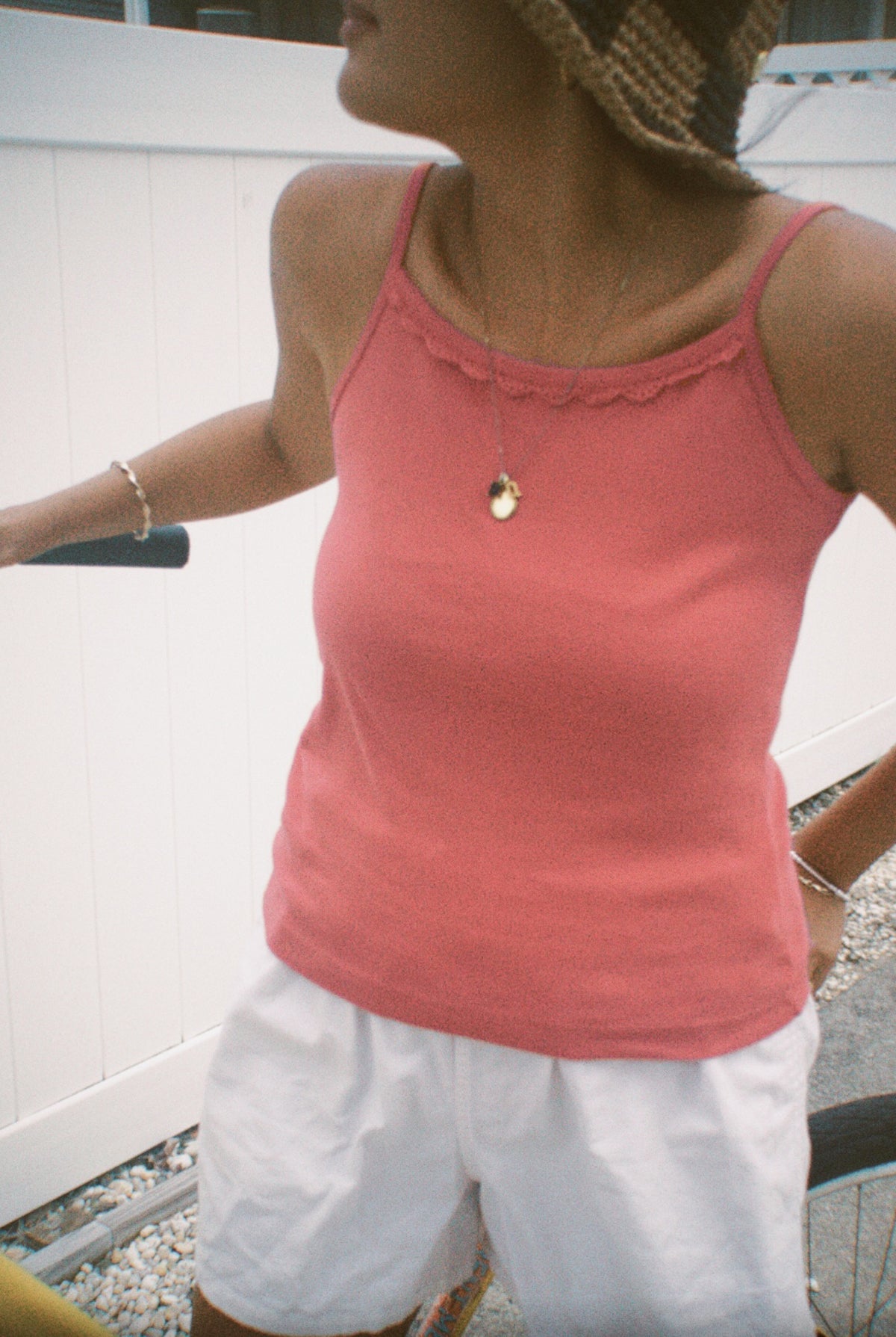 Rose -
A woman wears the LA RELAXED Frill Camisole in pink with white shorts, a straw hat, and jewelry, standing by a bicycle near a white fence on gravel. Her face is partially out of frame.