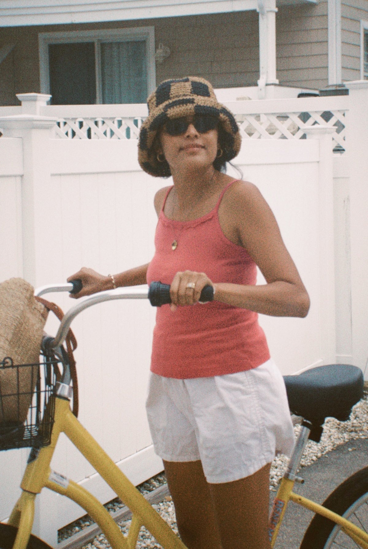 Rose -
A woman wearing the LA RELAXED Frill Camisole, white shorts, sunglasses, and a checkered bucket hat holds a yellow bicycle with a woven bag in its basket, posing in front of a white fence and house.