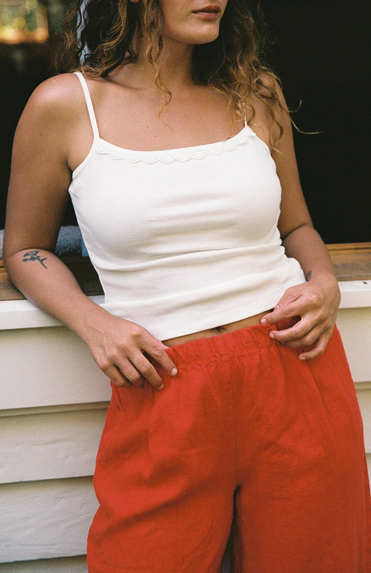 Cream -
A woman with wavy hair wears the LA RELAXED Frill Camisole, a white lace-trim top, paired with loose red pants and stands in front of a white wall, her hands on her waistband. Only her upper body is visible.
