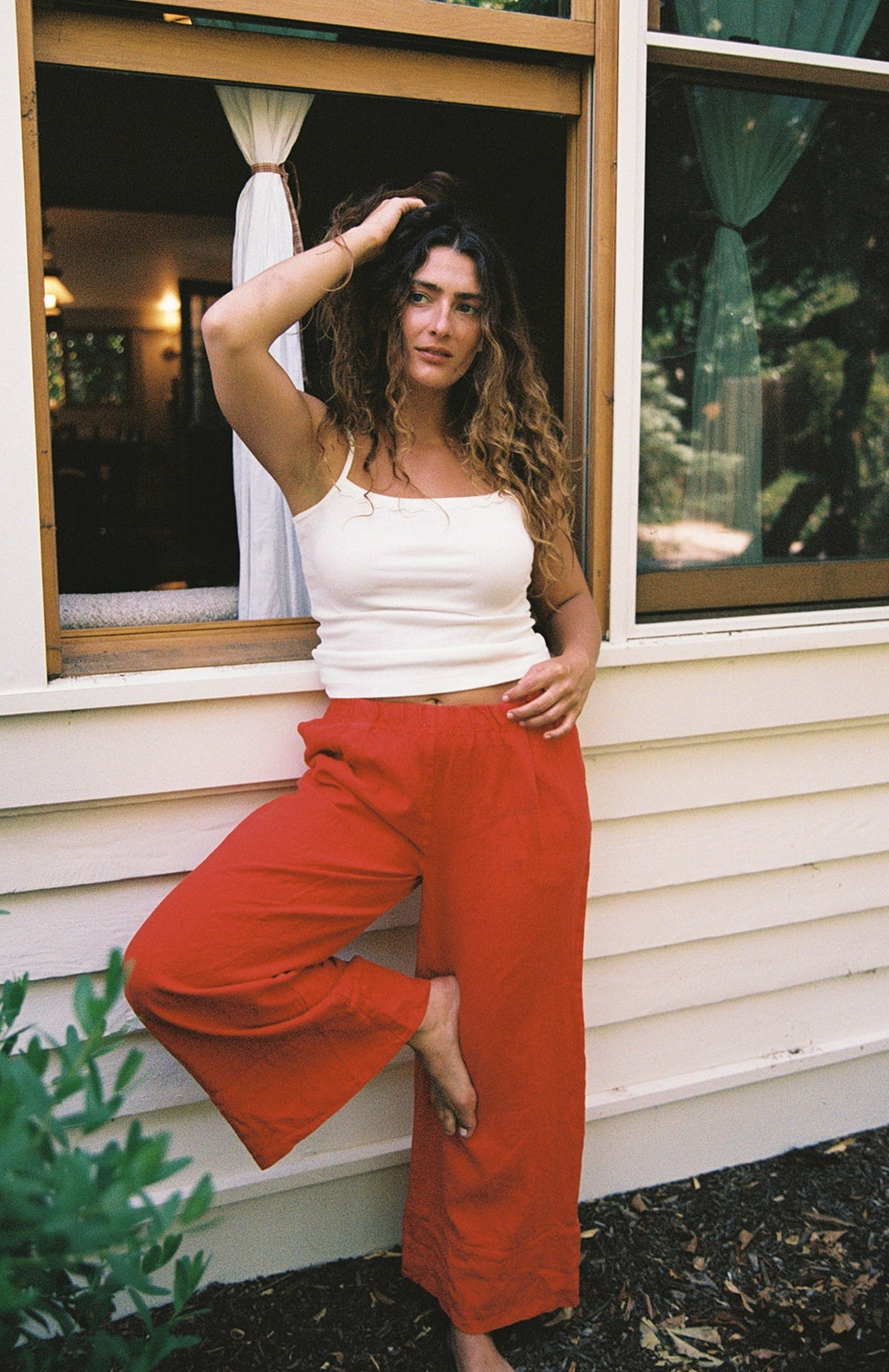 Cream -
A woman with curly hair wears the LA RELAXED Frill Camisole and red pants, standing barefoot by an open window with one leg bent, her hand in her hair as she looks at the camera.