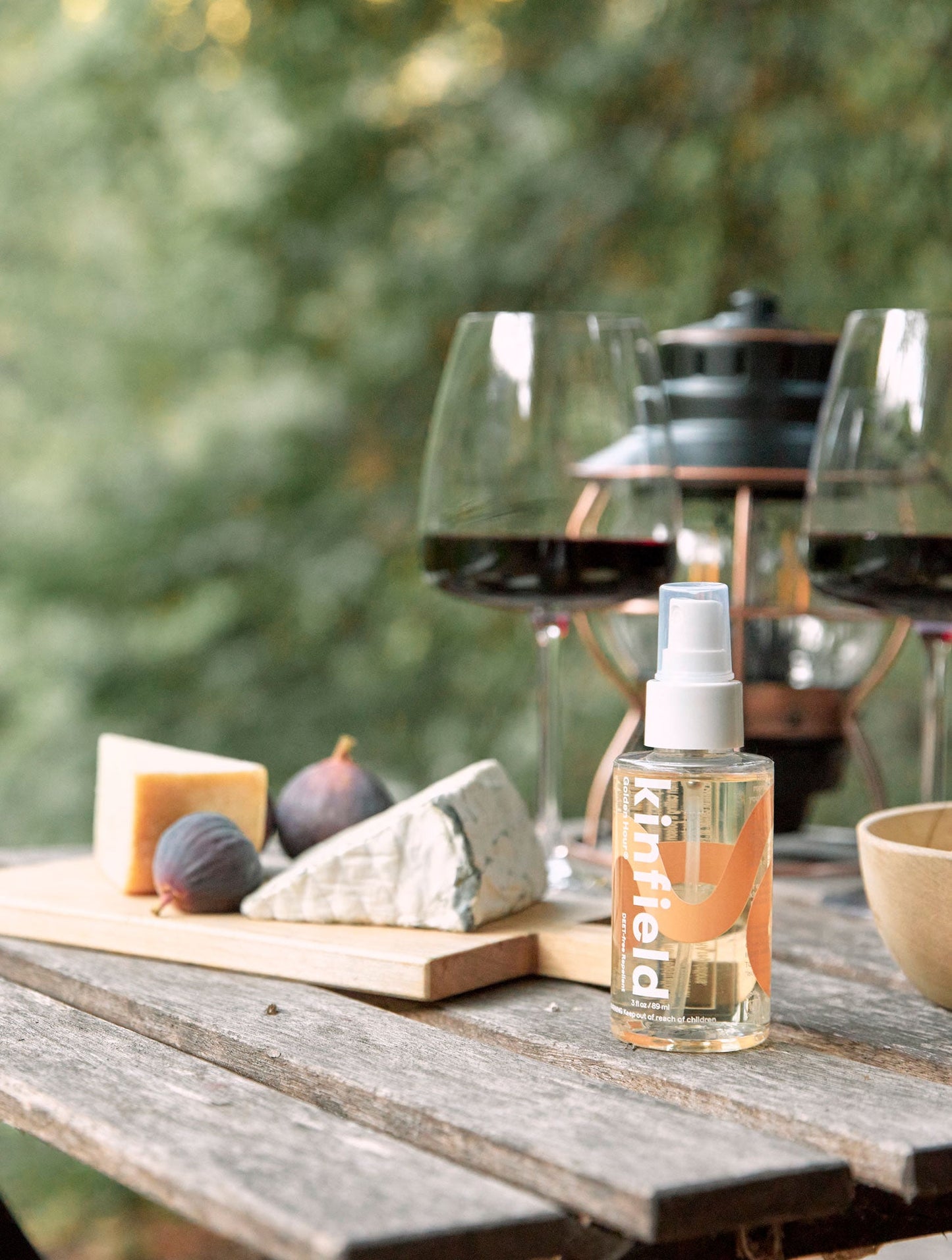 On a wooden outdoor table, there is a bottle of Kinfield's Golden Hour™ bug spray, accompanied by a cheese board with an assortment of cheeses and figs, along with two glasses of red wine, set against a softly blurred green backdrop.