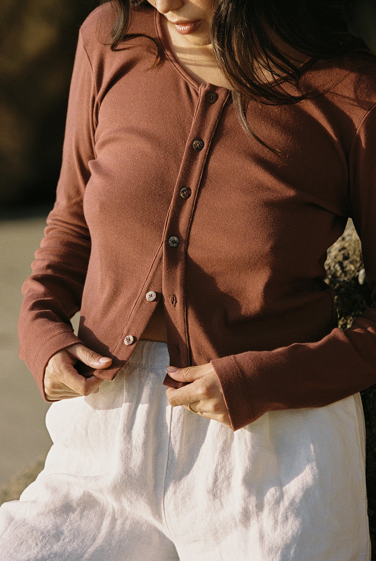 Someone is outdoors in natural sunlight, wearing an LA Relaxed Organic Rib Cardigan in rust over a white organic cotton top with white pants. They are adjusting the cardigan's buttons, creating a 90's closet vibe focused on the clothing.