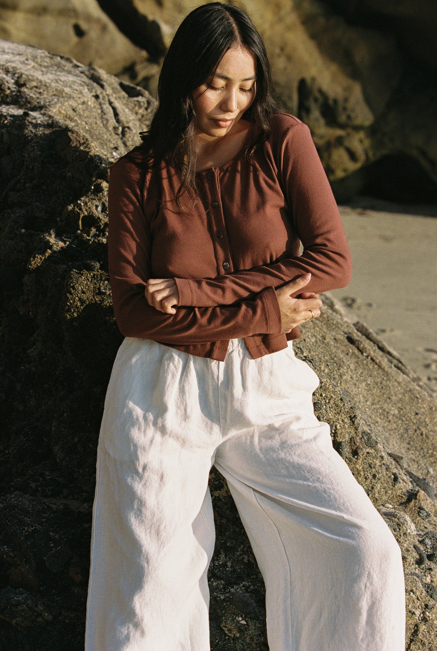 In a 90's Closet style, a person with long dark hair stands outdoors against a rock. They wear an LA Relaxed Organic Rib Cardigan in brown and white wide-leg pants made from organic cotton. The rocky setting is illuminated by natural light, possibly indicating a beach backdrop.