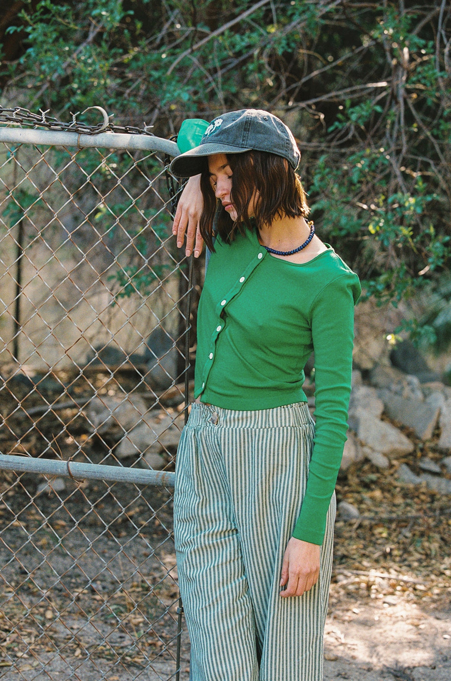 Someone wearing an Organic Rib Cardigan by LA Relaxed and striped pants leans on a chain-link fence outside. Sporting a dark hat with a '90s vibe, they exude relaxed style amidst greenery, effortlessly showcasing eco-friendly fashion made from organic cotton.