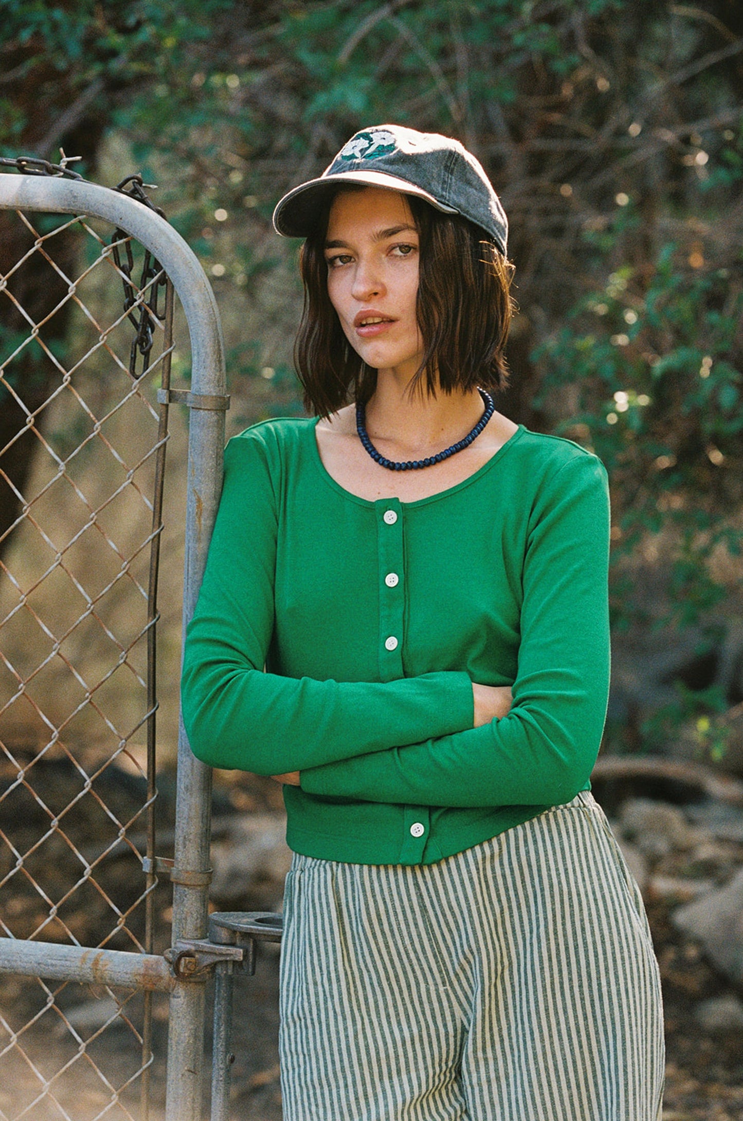 A person stands with arms crossed by a wire fence outdoors, wearing an LA Relaxed Organic Rib Cardigan, striped pants, and a denim cap. The lush greenery and soft lighting create a 90's closet vibe in the background.