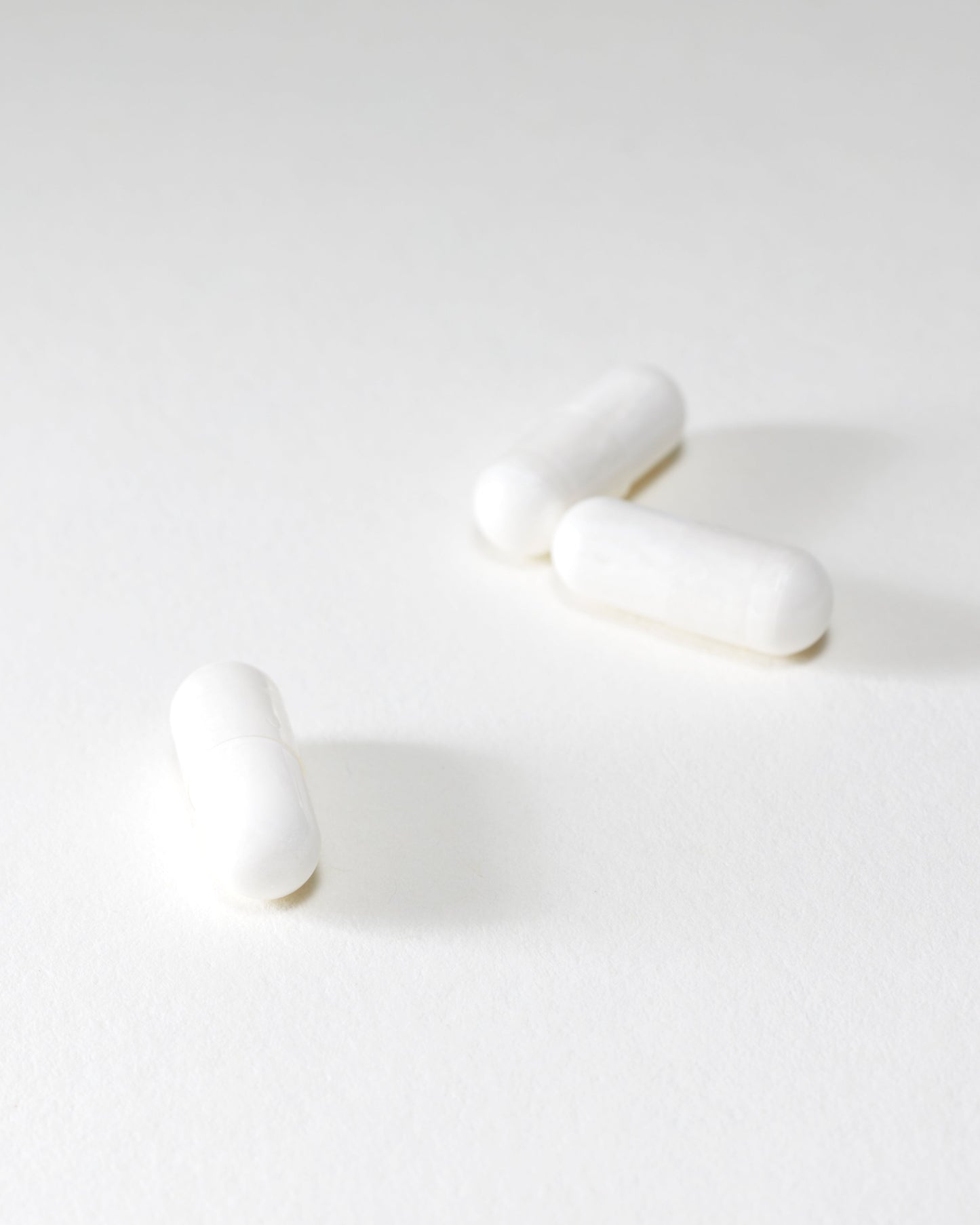 Three DOSAGE Mother-of-Pearl capsules are scattered on a pristine white surface. The image boasts a minimalistic and bright aesthetic, emphasizing the simplicity and uniformity of the capsules. Faint shadows add depth to the otherwise plain background.
