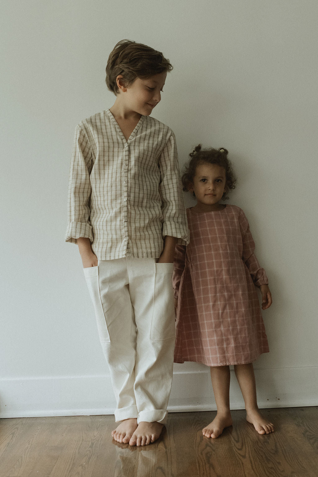 A young girl stands barefoot indoors against a plain wall wearing the TAKE CARE TOP olive check by cabane childrenswear, looking calm and relaxed.