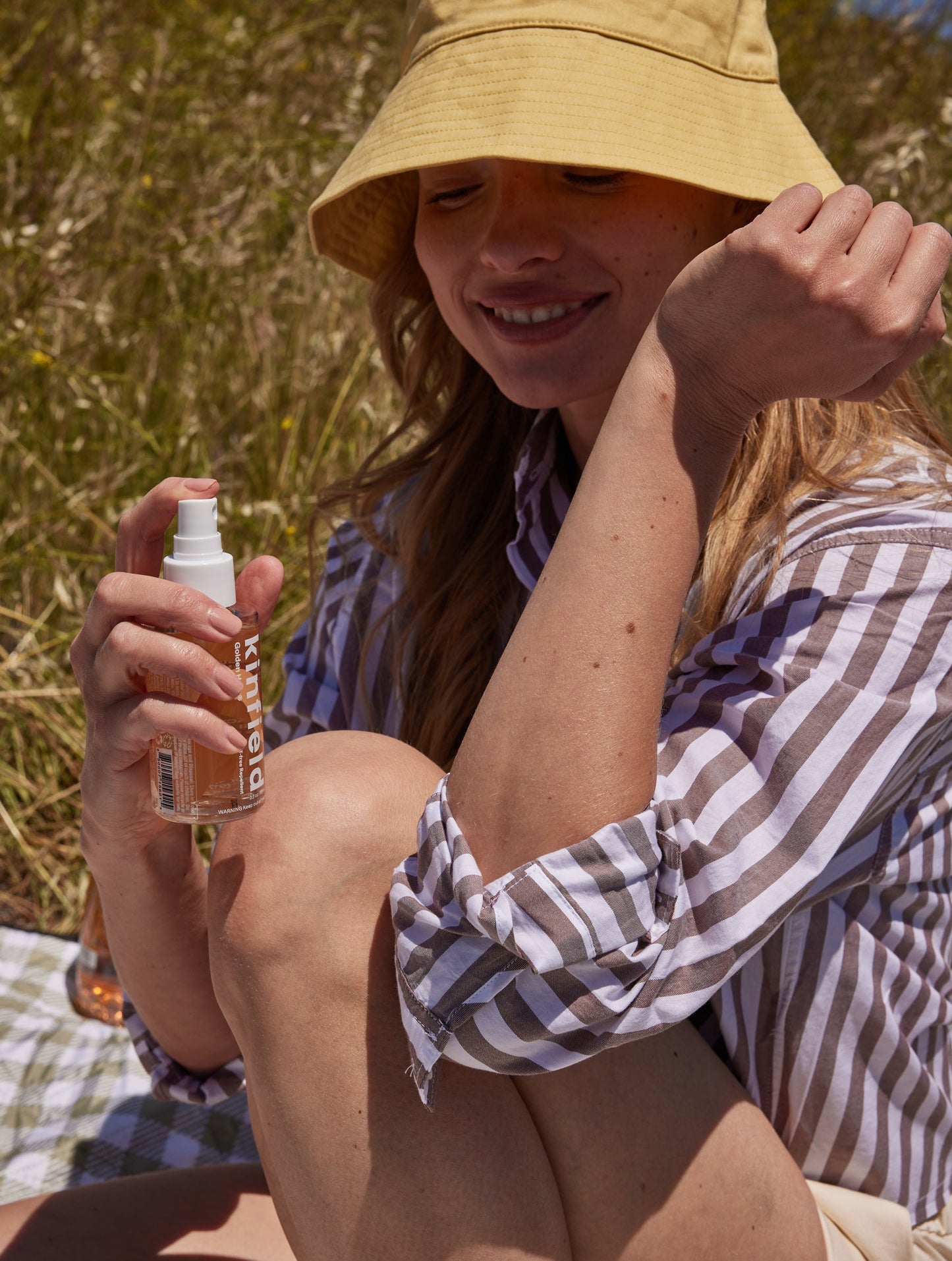 A person wearing a striped shirt and yellow bucket hat sits outdoors on a picnic blanket, holding Kinfield's Golden Hour™ spray bottle, which contains Indonesian citronella. They smile softly, surrounded by lush greenery and sunlight, relishing the moment while keeping bugs at bay.