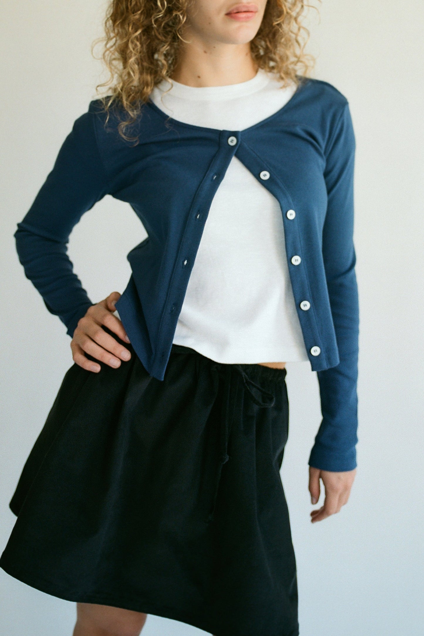 Channeling a 90's Closet vibe, a model stands with hand on hip wearing LA Relaxed's blue Organic Rib Cardigan over a white top and black skirt. The cardigan is stylishly half-unbuttoned, showcasing its organic cotton fabric against a plain light backdrop. Curly hair completes the look.