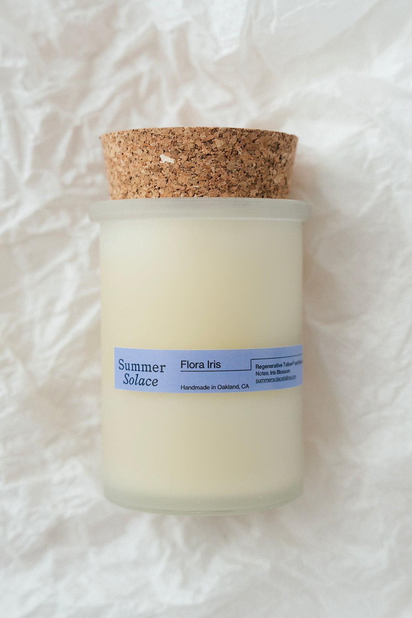 A frosted glass jar candle with a cork lid sits on crumpled white paper. The blue label reads "Summer Solace Tallow" featuring the "Flora Iris Candle - Regenerative Tallow®," enhanced with orris butter, and notes that it's handmade, offering a unique aromatic experience.