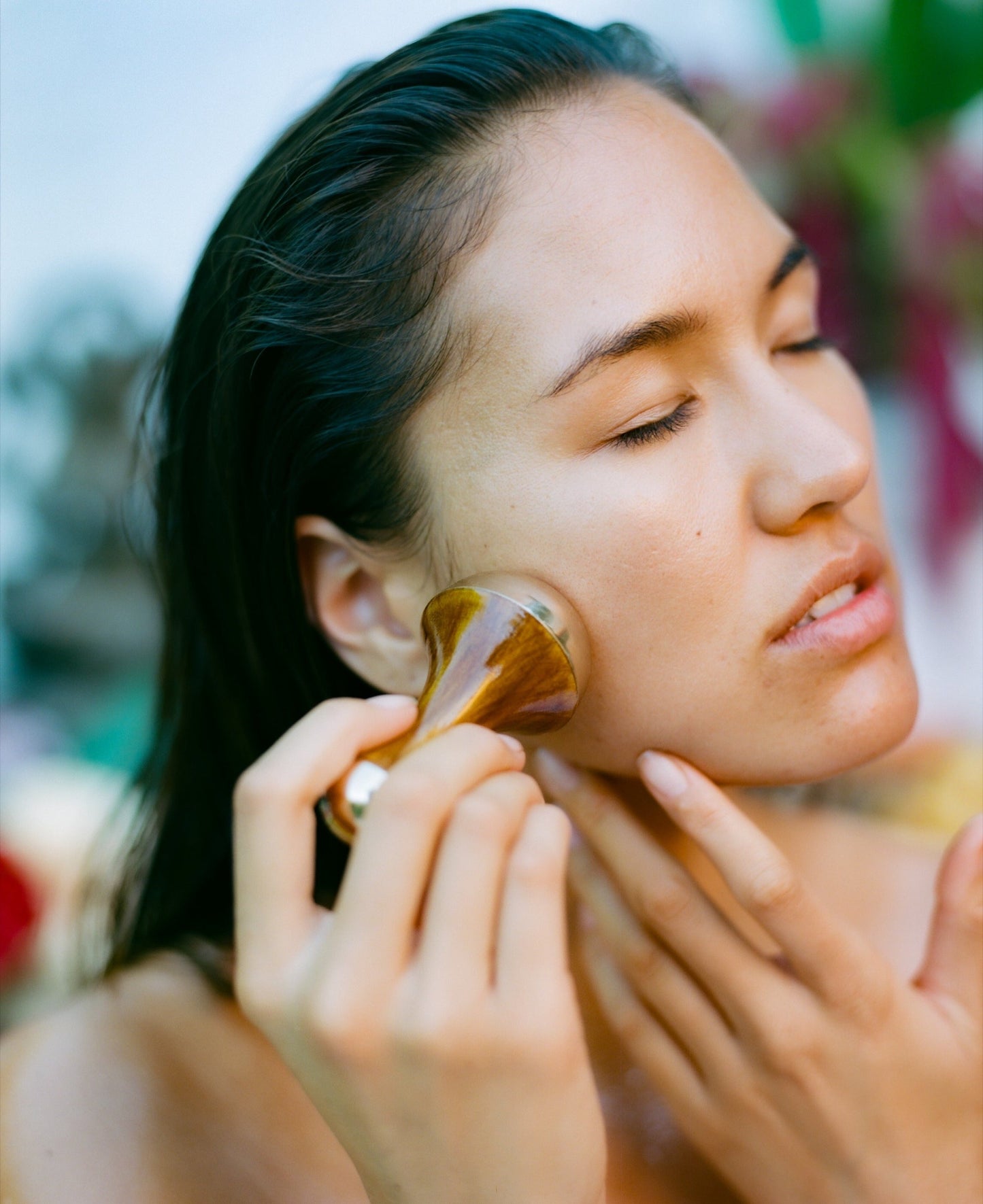 A person with wet hair is shown up close, eyes closed, and gently massaging their cheek with a Surya Kansa Wand. The background is blurred with hints of greenery and colorful elements, highlighting the serene moment with this Ayurvedic skincare tool.
