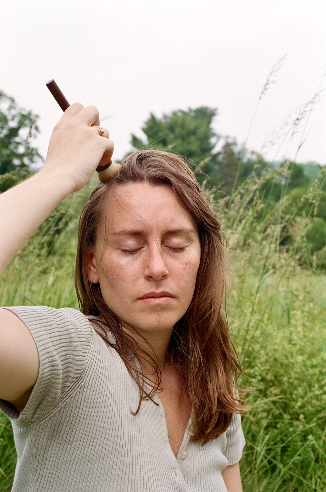 A woman with long natural hair and closed eyes stands in a grassy field, lifting the & Tread Well Dry Shampoo Brush above her head as if brushing her hair. She wears a light short-sleeve shirt and is surrounded by lush greenery.