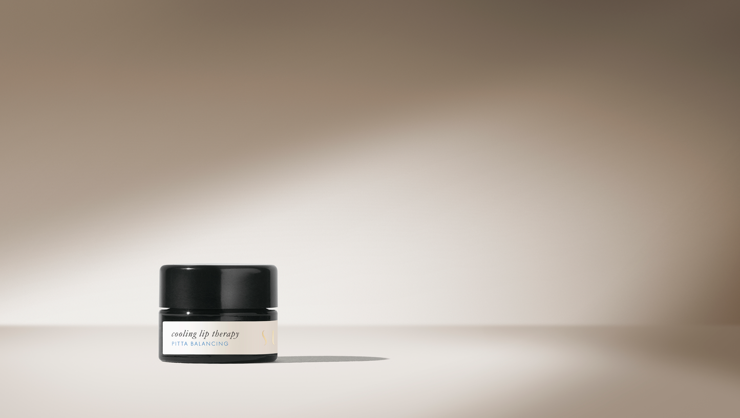 A small black container of Surya's "Cooling Lip Therapy" labeled "Pitta Balancing" sits on a smooth beige surface. Featuring organic ingredients, the backdrop is softly lit, highlighting the natural blend with a gentle gradient of shadows and light.