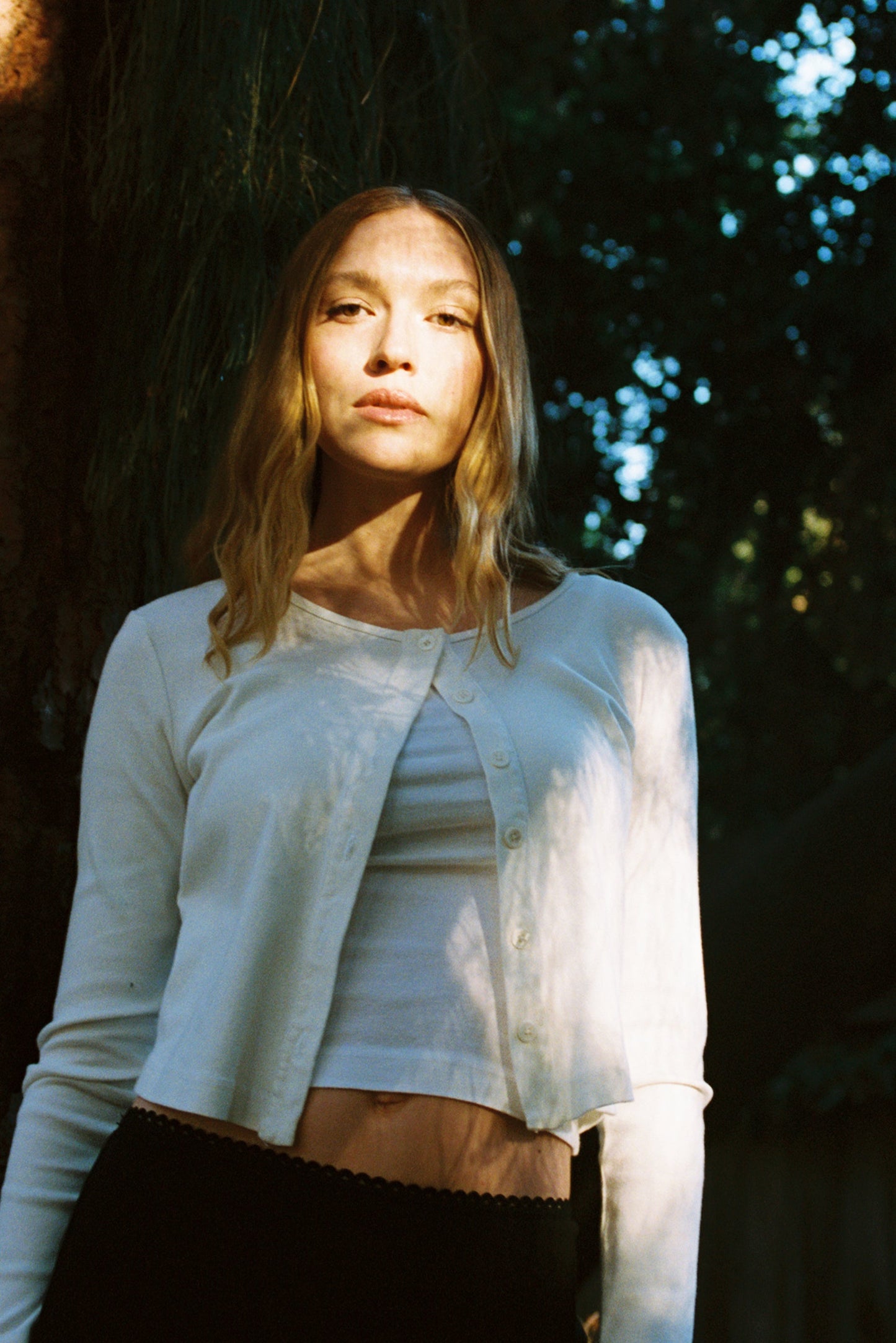 A woman with long, wavy hair stands in a forest wearing an LA Relaxed Organic Rib Cardigan over a white top. Sunlight filters through the trees, creating patterns on her as she gazes calmly at the camera, embodying a serene 90s closet vibe.