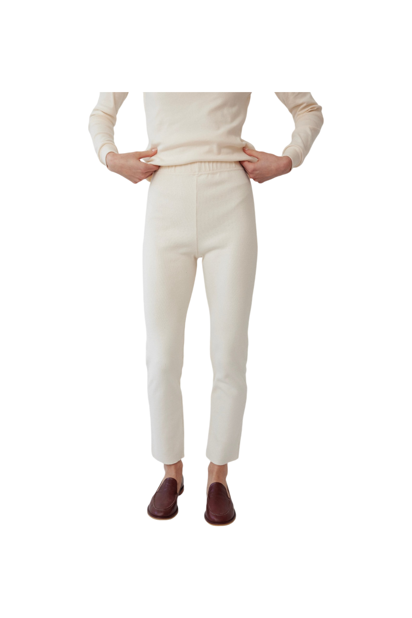 The person is dressed in a cream long-sleeve top and fitted Easy Legging: Natural from Wol Hide, with their hands on their waist. The leggings are made from stretchy rib fabric, and they complement maroon slip-on shoes. The scene is set against a plain white background.