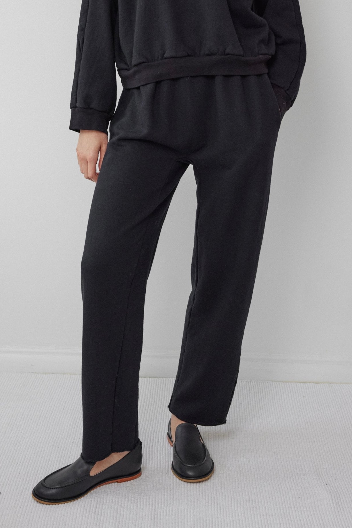 A person is standing on a light-colored carpet, dressed in the Wol Hide Easy Winter Sweatshirt in black and matching trousers. Their outfit, including the sleek black slip-on shoes, complements the plain white wall backdrop. The sweatshirt's organic cotton fleece offers comfort and is proudly made in the USA.