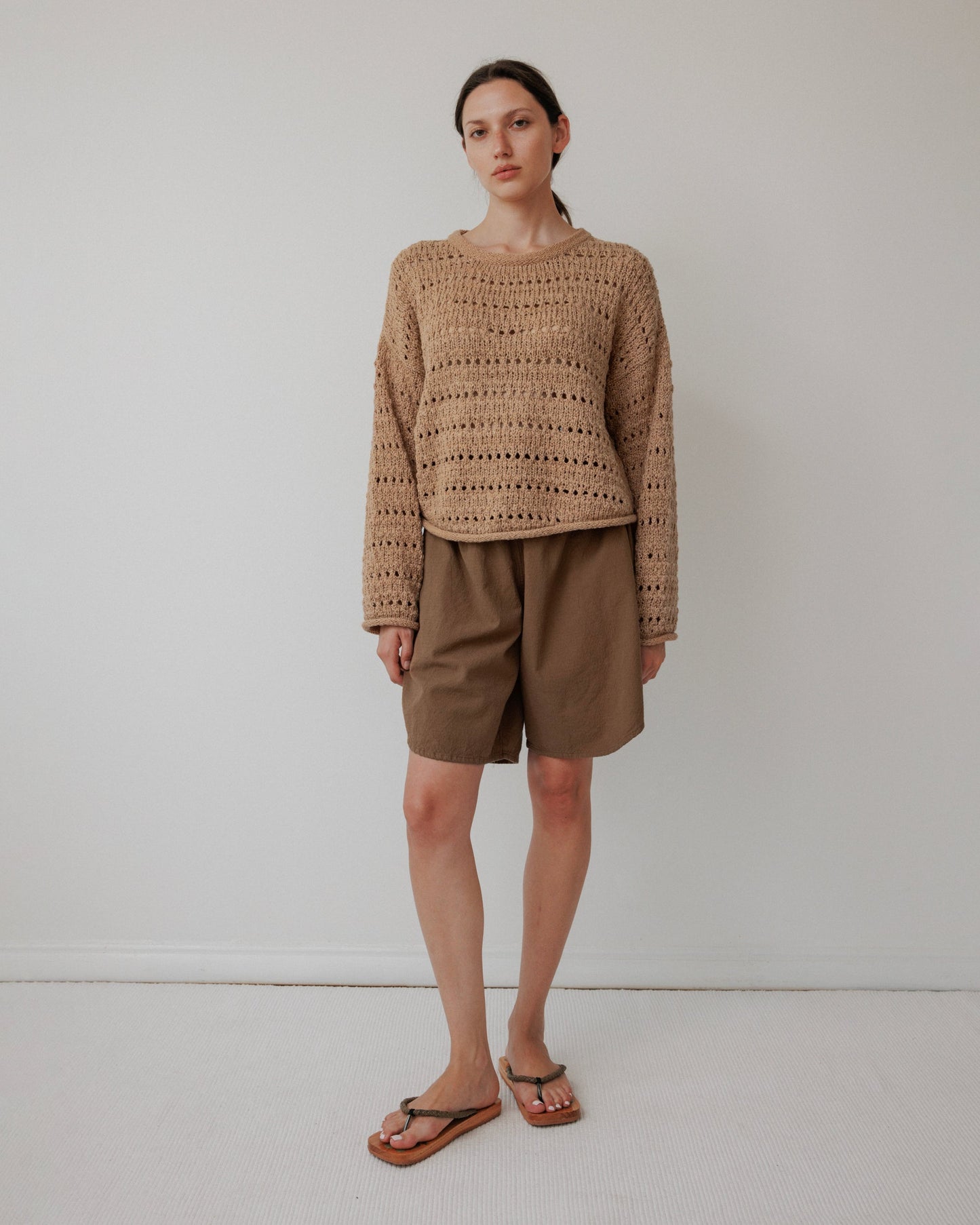 Someone stands against a light wall, wearing an Open Pullover : Tannin from Wol Hide, paired with khaki shorts and black flip-flops. Their hair is pulled back as they gaze neutrally at the camera.