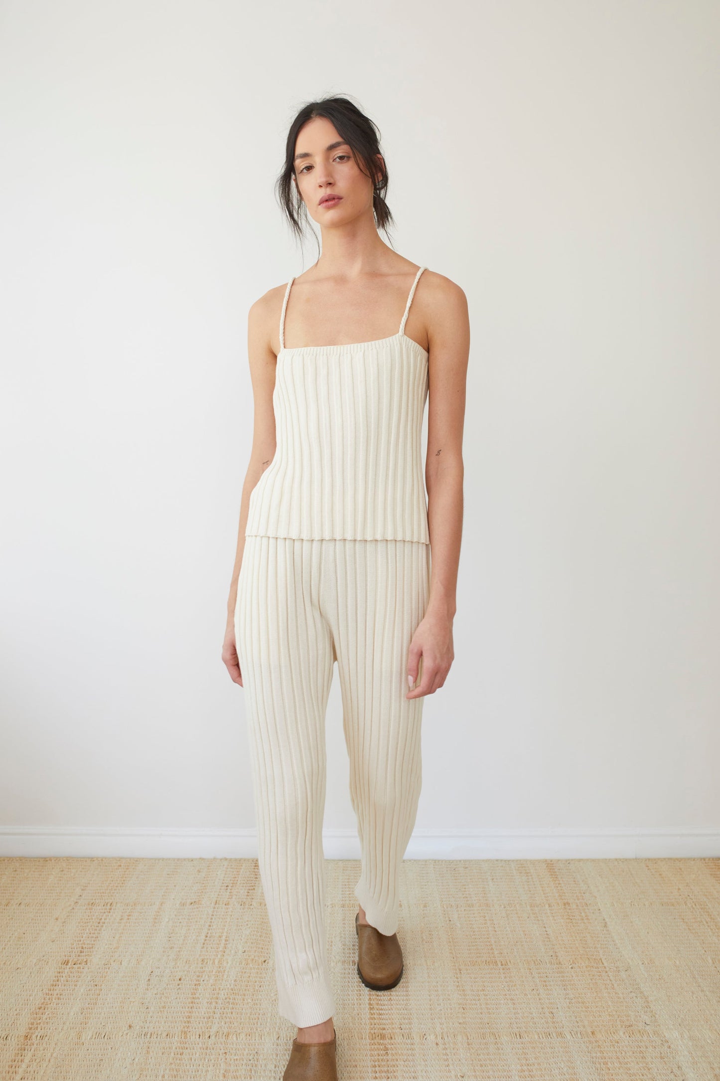 A person stands against a plain white wall, wearing the Wol Hide Rib Cami in Ecru—a cream-colored ribbed tank top with braided straps—and matching pants made from regenerative cotton. They have long, dark hair tied back and wear tan shoes. The floor is hardwood.
