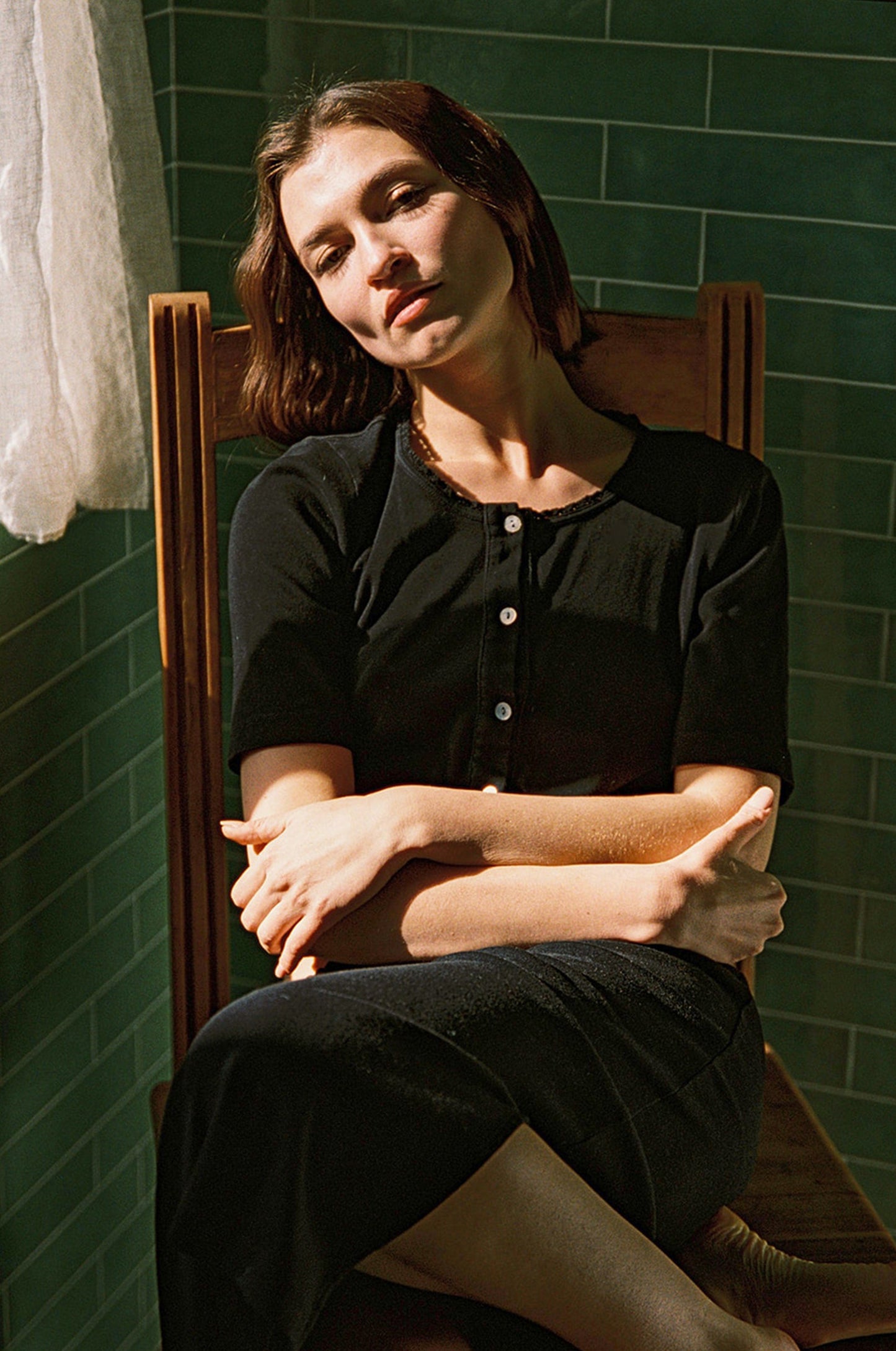 A woman in a Short Sleeve Organic Cardi by LA Relaxed sits on a wooden chair, arms crossed and eyes closed, bathed in soft sunlight. The green tiled walls and white curtain form a serene backdrop.