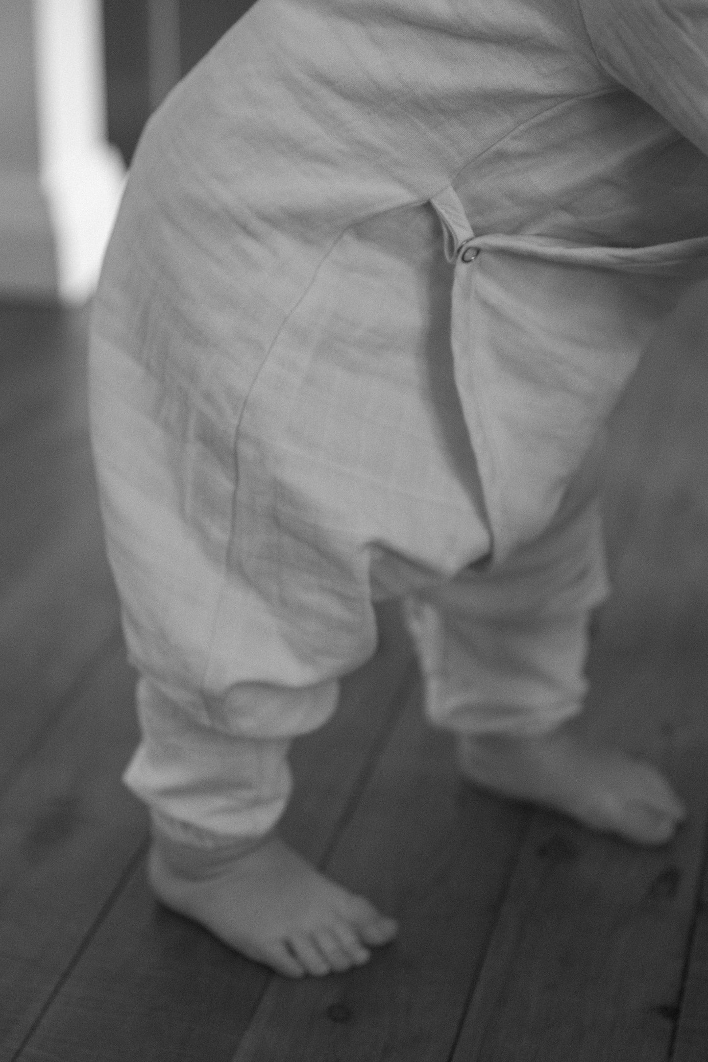 A black and white image captures the lower half of a small child, barefoot on a wooden floor, wearing a loose, light-colored apres wrap from breathable fabric.