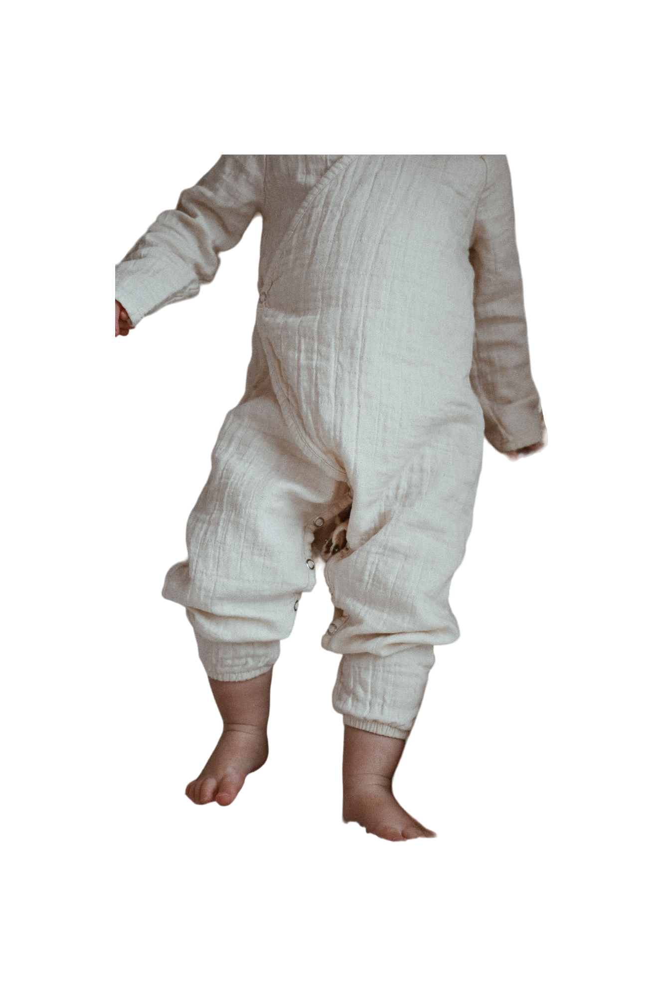 A baby stands wearing the Après Organic Wrap Undyed onesie by cabane childrenswear, crafted from breathable fabric in a soft, natural hue. With bare feet, the baby appears to be either taking small steps or standing still.