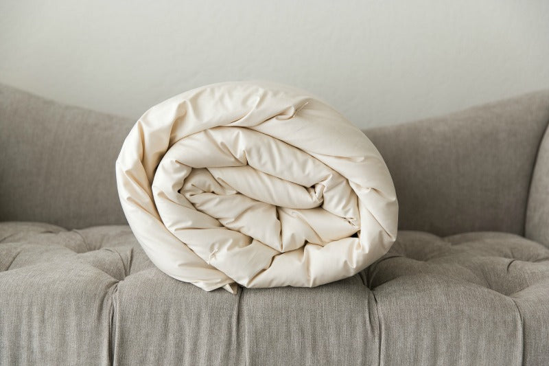 The Ashland Organic Wool Comforter from Shepherd's Dream, elegantly rolled in beige, rests on a tufted gray sofa. The image showcases the texture and folds of the comforter against a clean, minimalist background.