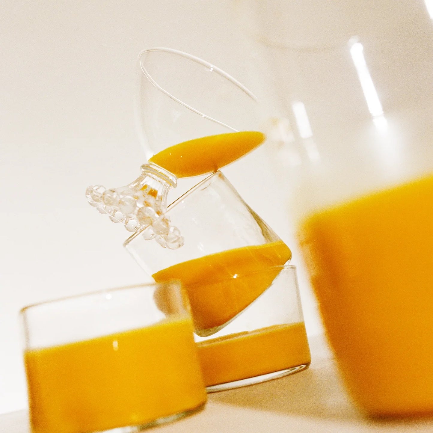 A close-up of three glasses tilted and stacked, each containing Warm Feelings 100-Day Supply by THE FULLEST, shows the vibrant hues reminiscent of a saffron latte. The dynamic arrangement against a neutral background highlights the soothing essence of the orange juice.