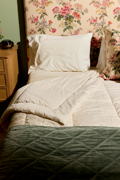 A large, puffy white comforter is draped over a thick tree branch in a lush, sunlit forest clearing. The scene appears tranquil and serene with natural light filtering through the trees.