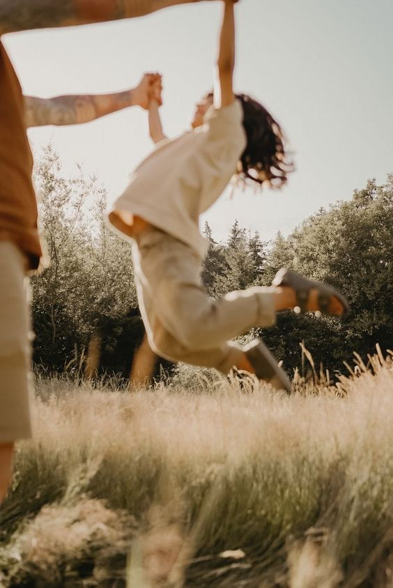 A child is joyfully swinging while holding onto an adult's hand in a sunlit grassy field. The child is airborne, with legs bent and hair flying, capturing a moment of playful happiness. Trees and a clear sky are in the background.