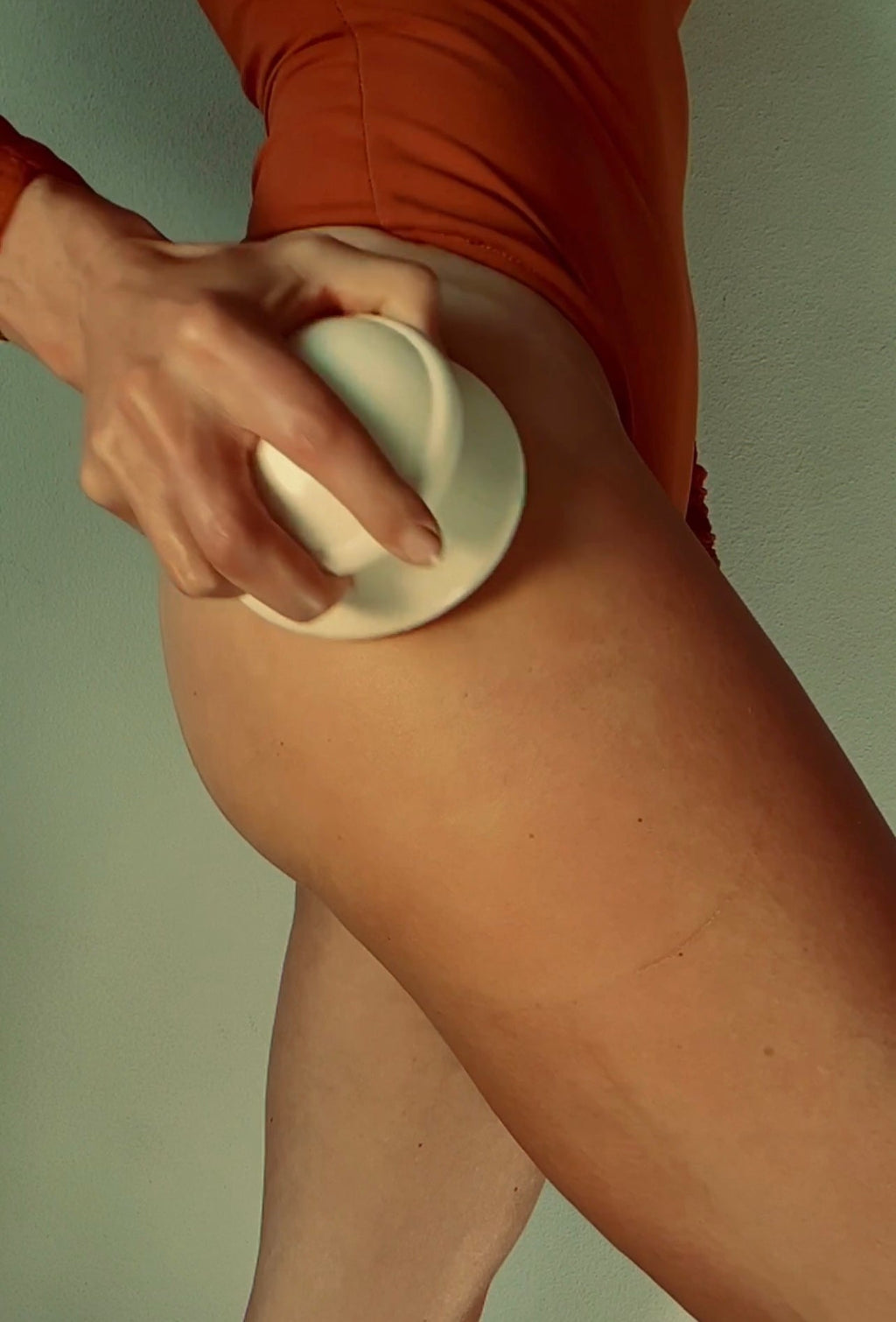 Wearing an orange swimsuit, a person uses the LENI THE FACE & BODY CUPPING SET—white lymphatic drainage cups—on their upper thigh, against a plain background.