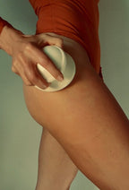 Wearing an orange swimsuit, a person uses the LENI THE FACE & BODY CUPPING SET—white lymphatic drainage cups—on their upper thigh, against a plain background.