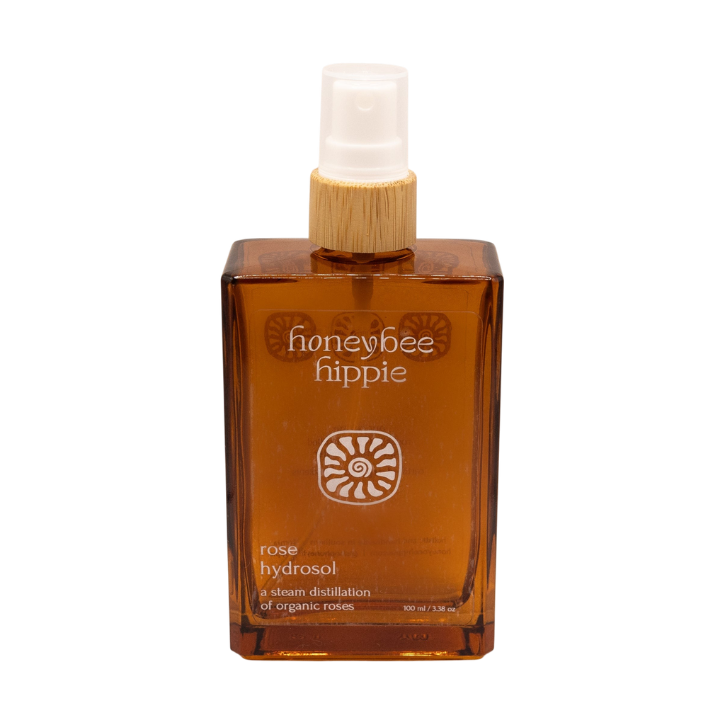 A brown rectangular spray bottle labeled "Honeybee Hippie rose hydrosol" with white text, sun-like symbols, a white spray nozzle, and a wooden collar—ideal as a natural toner or skin refresher.
