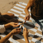 Four friends relax on a striped picnic blanket at the beach, each holding a tube of Honeybee Hippie vanilla & chamomile tallow lip butter and joining them together in the center of their group.