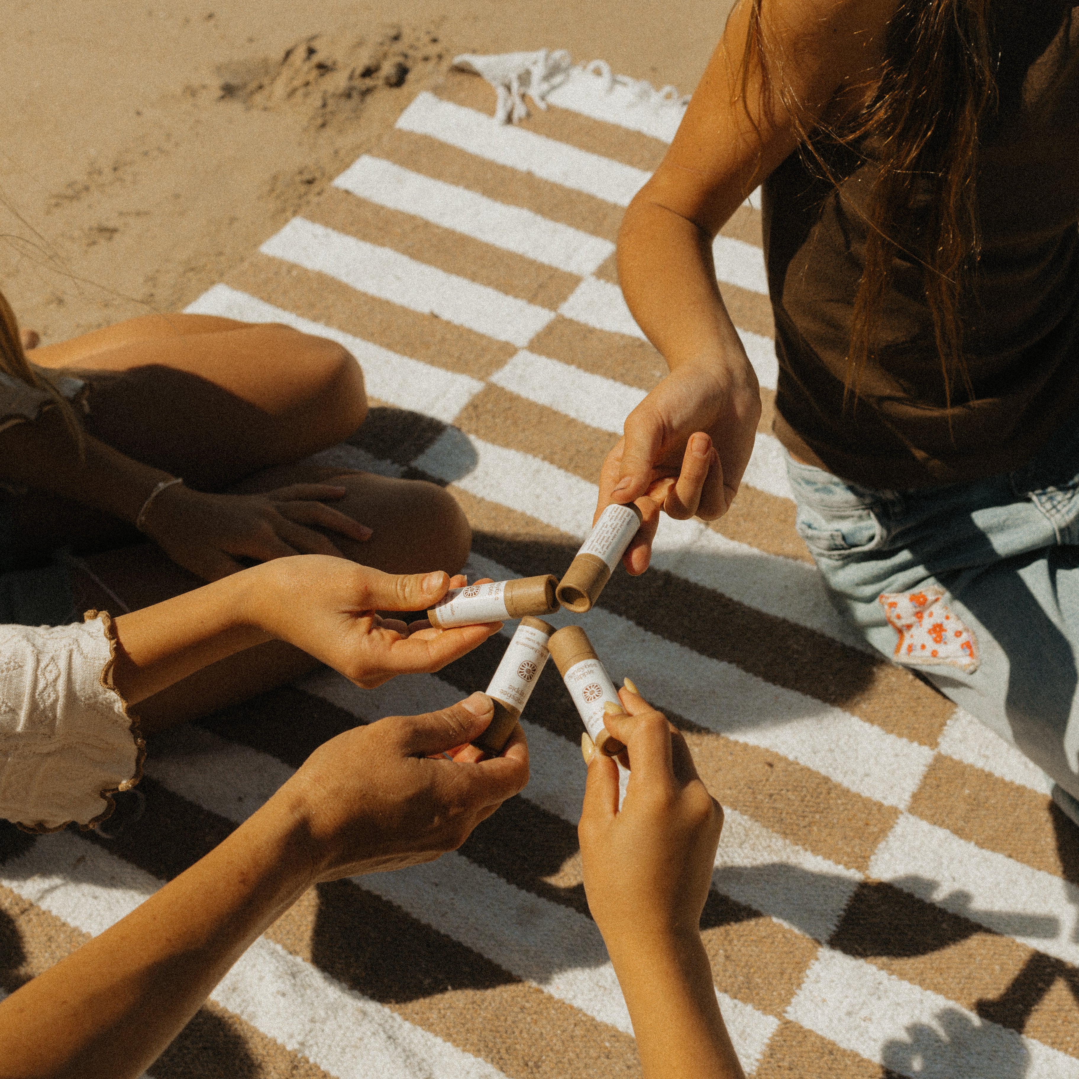 Four friends relax on a striped picnic blanket at the beach, each holding a tube of Honeybee Hippie vanilla & chamomile tallow lip butter and joining them together in the center of their group.