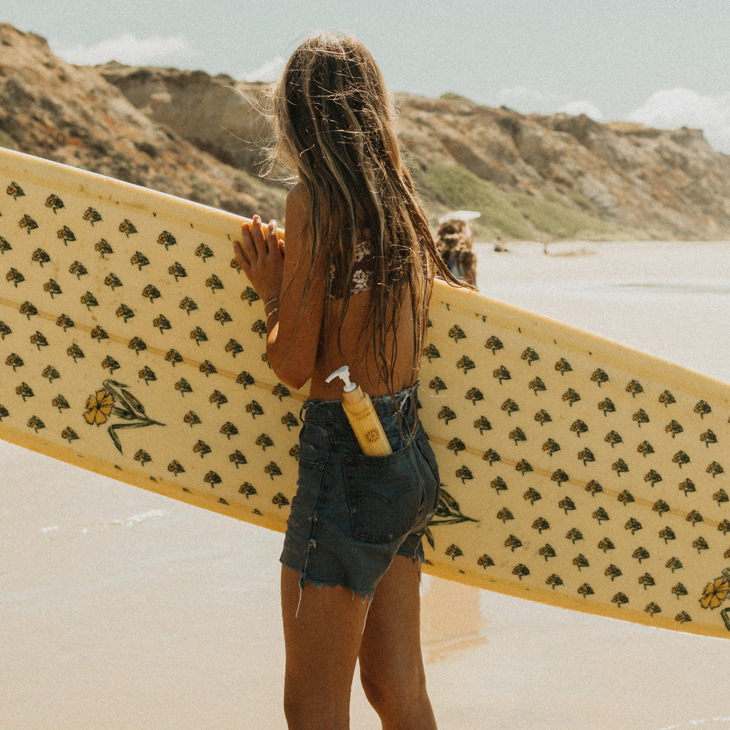 Beneath rocky cliffs and a blue sky, a sun-kissed woman in a bikini top and denim shorts tucks Honeybee Hippie’s california sunshine sunscreen into her pocket, carrying her patterned surfboard along the sunny beach.