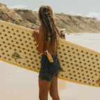 Beneath rocky cliffs and a blue sky, a sun-kissed woman in a bikini top and denim shorts tucks Honeybee Hippie’s california sunshine sunscreen into her pocket, carrying her patterned surfboard along the sunny beach.