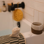 A close-up of a bathtub with a black faucet, yellow sponge, striped towel, and Honeybee Hippie postpartum herbal sitz bath in a linen pouch for soothing postpartum care. A ceramic cup of tea completes this warm, inviting scene.