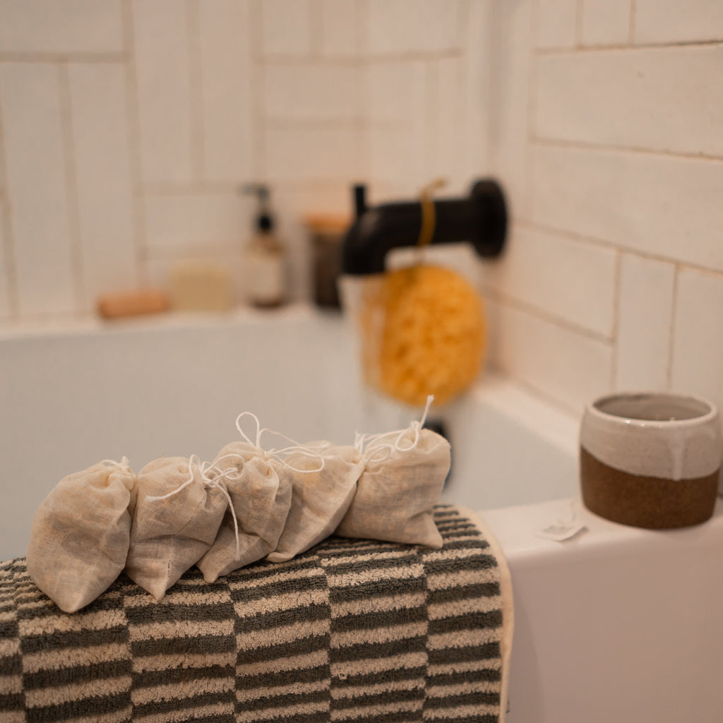 Four Honeybee Hippie postpartum herbal sitz bath pouches rest on a striped towel by the tub, with a sponge, faucet, and ceramic cup nearby against white tiles—creating a calming self-care scene.