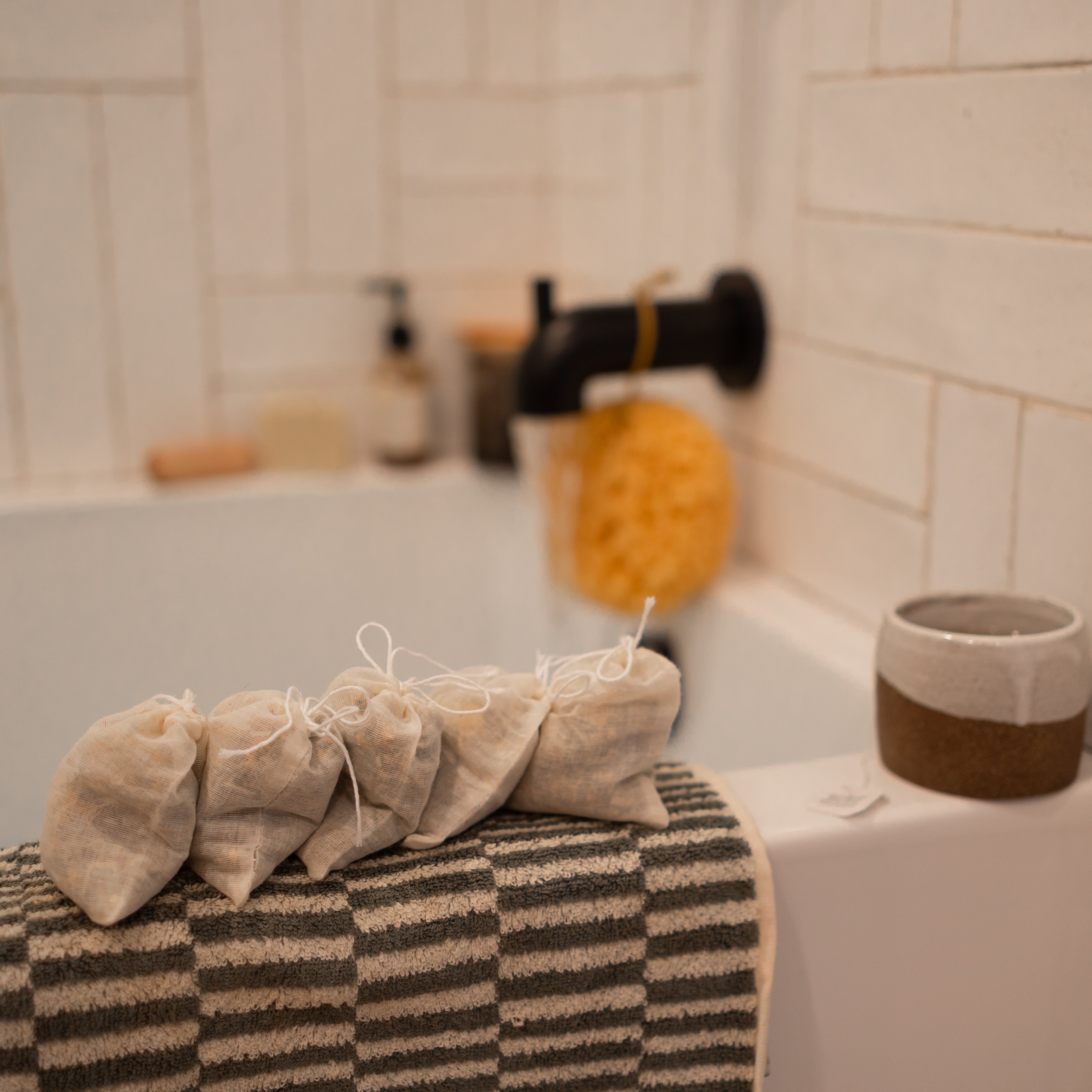 Four Honeybee Hippie postpartum herbal sitz bath pouches rest on a striped towel by the tub, with a sponge, faucet, and ceramic cup nearby against white tiles—creating a calming self-care scene.