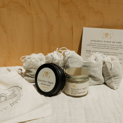 The Honeybee Hippie's natural mama bundle includes a black tin labeled "miracle mud," a jar of tallow called "belly balm," fabric bags, and a note card with sitz bath instructions, all set on a wooden background.