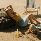 A woman sporting sunglasses and glowing skin lounges by a rock on the sand as a toddler plays nearby. Next to them are a striped beach chair, Honeybee Hippie’s california sunshine body oil, and sunscreen.