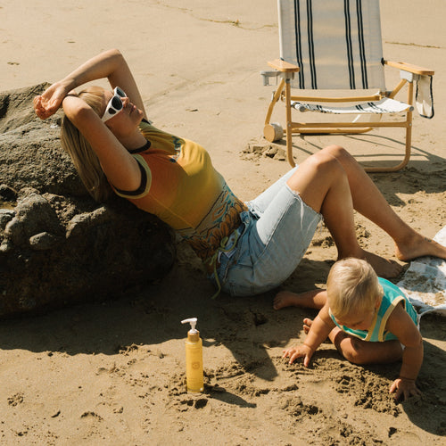 A woman sporting sunglasses and glowing skin lounges by a rock on the sand as a toddler plays nearby. Next to them are a striped beach chair, Honeybee Hippie’s california sunshine body oil, and sunscreen.