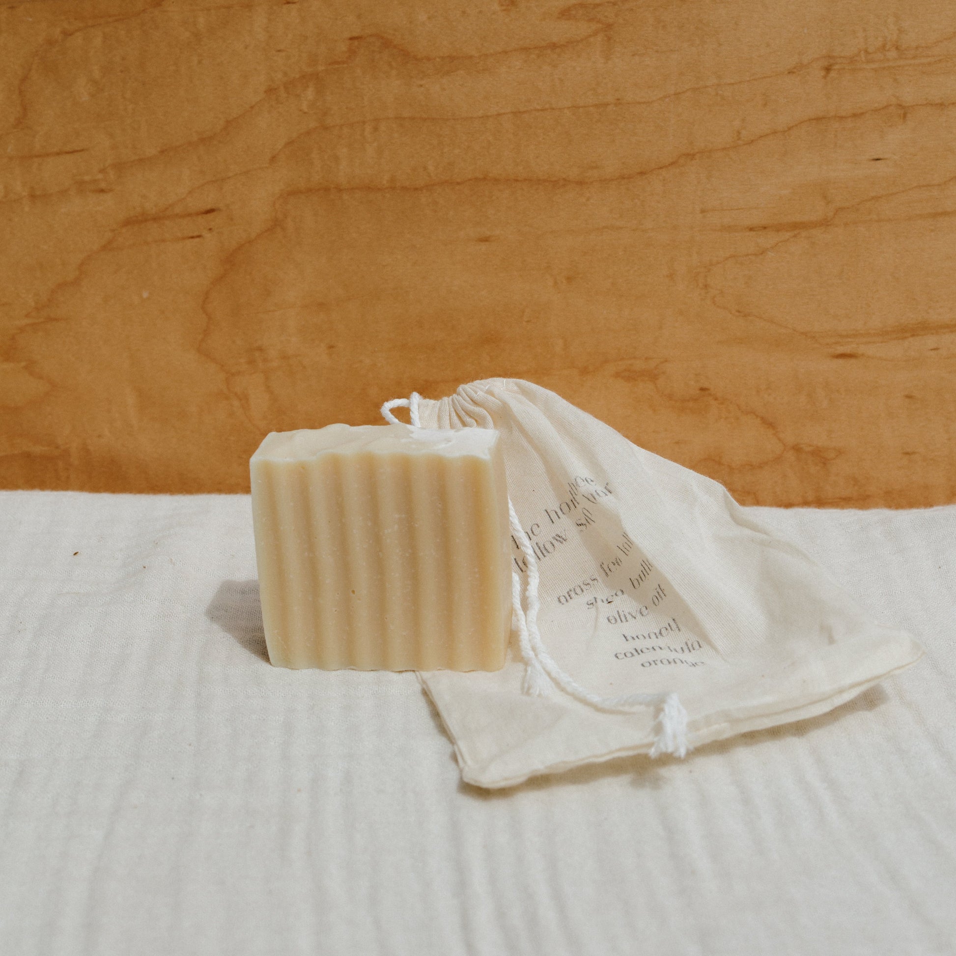 The Honeybee Hippie honeybee tallow soap bar, with its textured rectangular shape, sits against a light brown wooden background. It's in front of a cream-colored drawstring bag with subtly printed text, lying on a beige cloth surface.